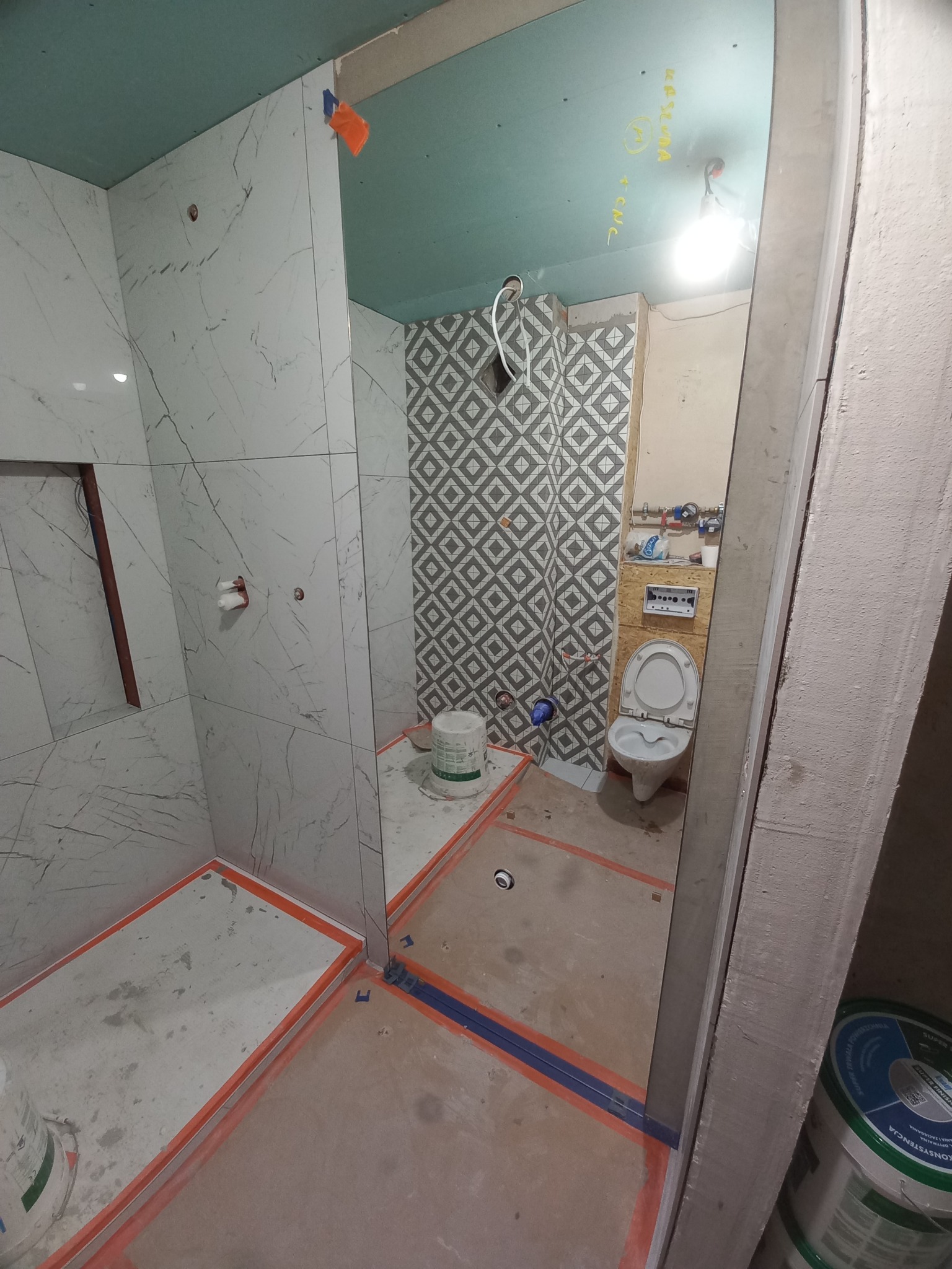 Wnętrze łazienki w trakcie remontu: białe płytki imitujące marmur na ścianach prysznica, geometryczne płytki na ścianie z toaletą, sufit z płyt kartonowo-gipsowych, widoczne instalacje...