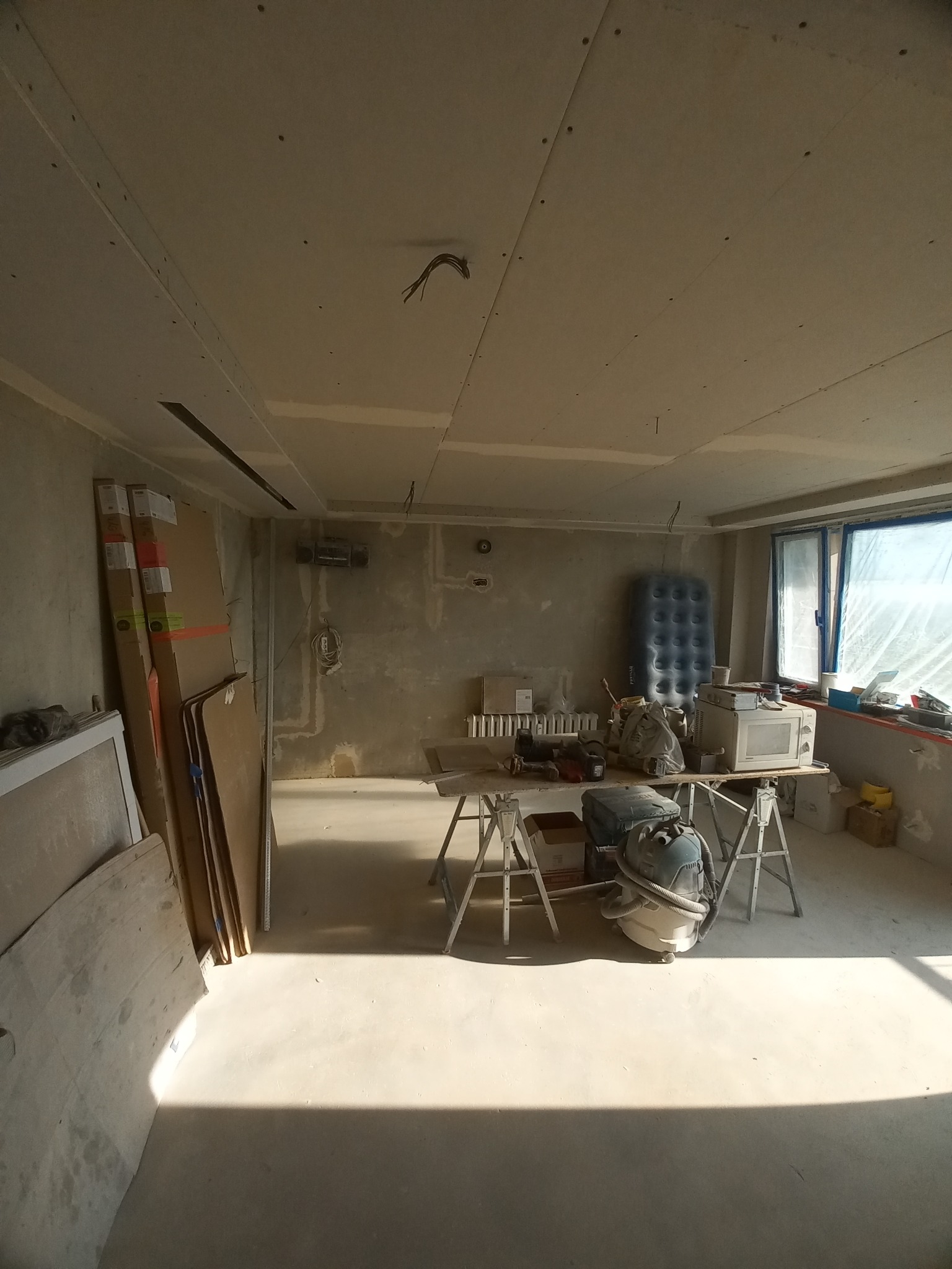 Wnętrze w trakcie prac wykończeniowych: surowe ściany, zamontowane płyty gipsowo-kartonowe na suficie, widoczne przewody elektryczne, narzędzia pracy i odkurzacz przemysłowy na tle okna zakrytego...