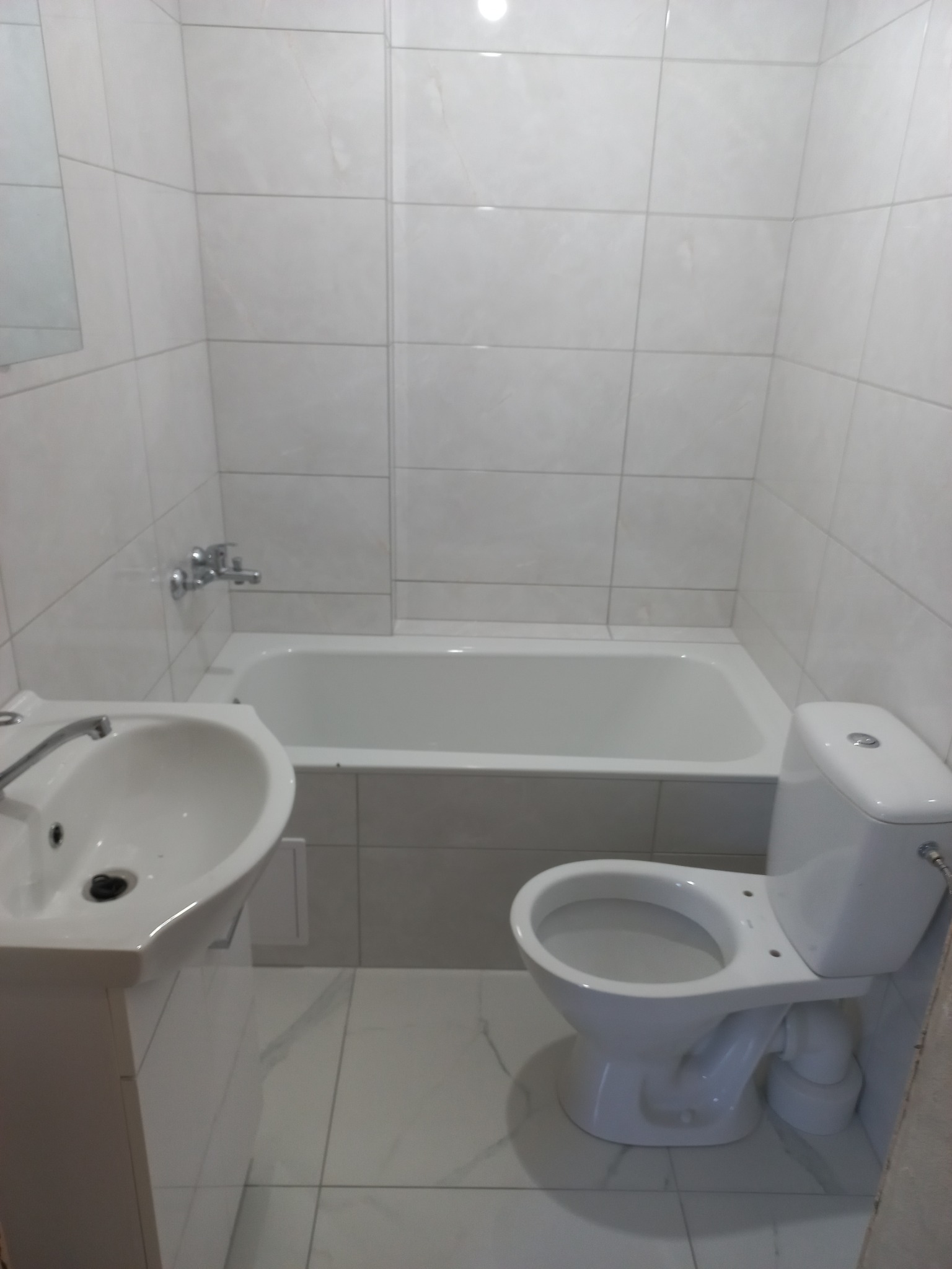 Wykończona łazienka z białą armaturą: umywalka na postumencie, toaleta kompaktowa i wanna obudowana płytkami, całość w jasnej tonacji.