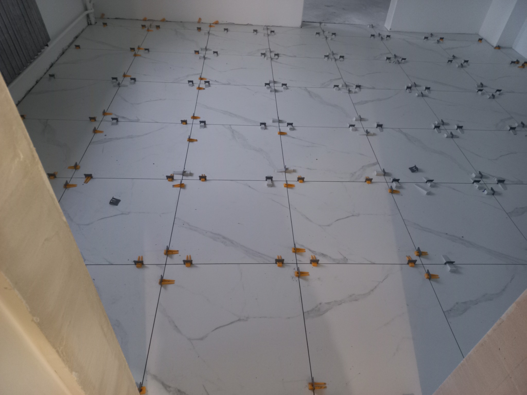 Układanie białych płytek podłogowych z marmurkowym wzorem, z użyciem czarnych krzyżyków dystansowych i pomarańczowych klinów poziomujących.