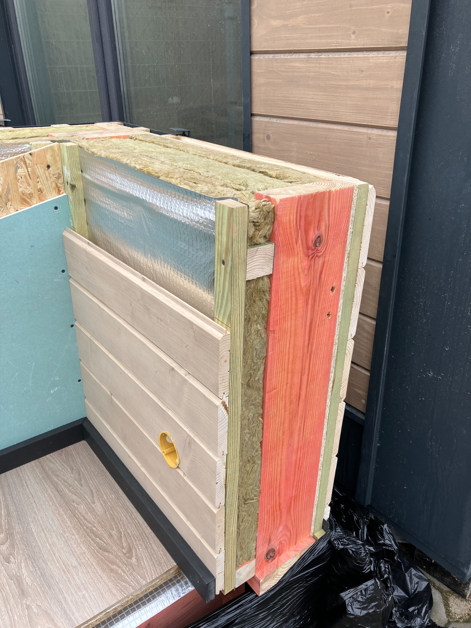 Przekrój ściany domu w trakcie budowy, ukazujący warstwy izolacji, folię aluminiową, drewniane deski elewacyjne z otworem na instalację elektryczną, oraz fragment podłogi.