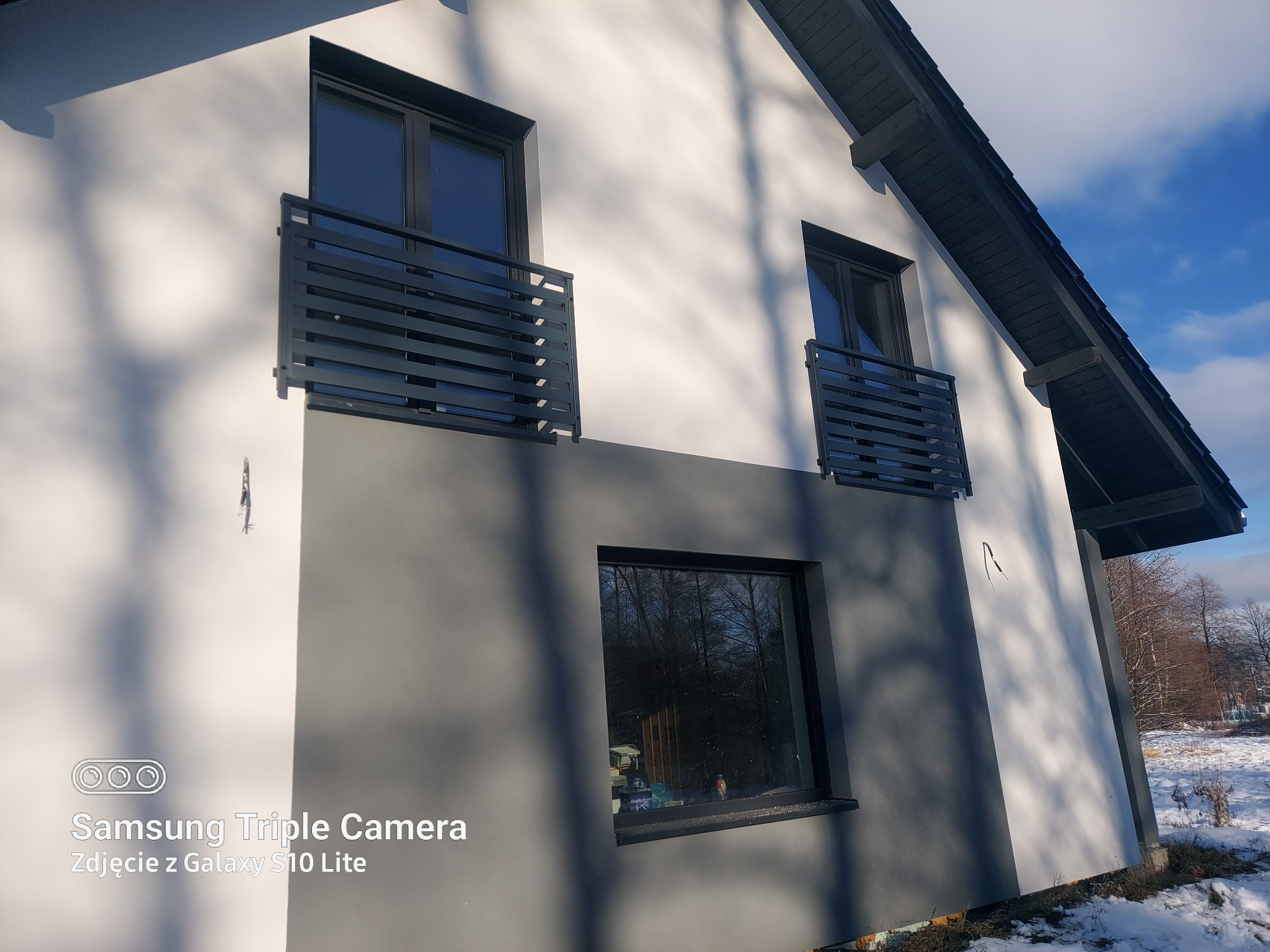 Dwukondygnacyjny dom z szarą i białą elewacją, czarnymi ramami okiennymi i balkonami z metalowymi balustradami, na tle zimowego krajobrazu i błękitnego nieba.