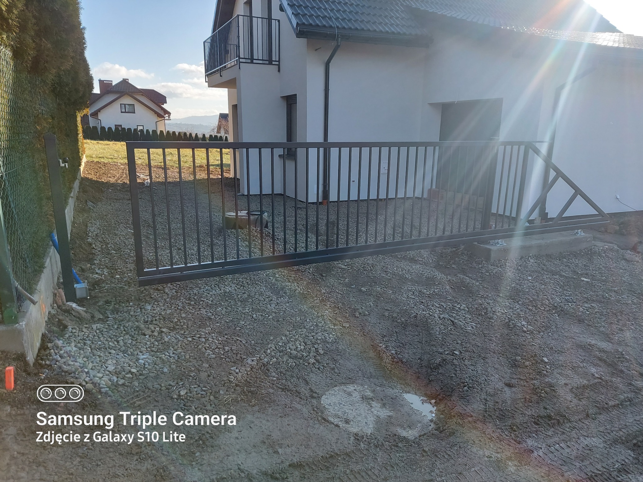 Nowa, przesuwna brama wjazdowa z metalowymi prętami na posesji z nowym domem, widoczne ślady prac ziemnych i gruz.