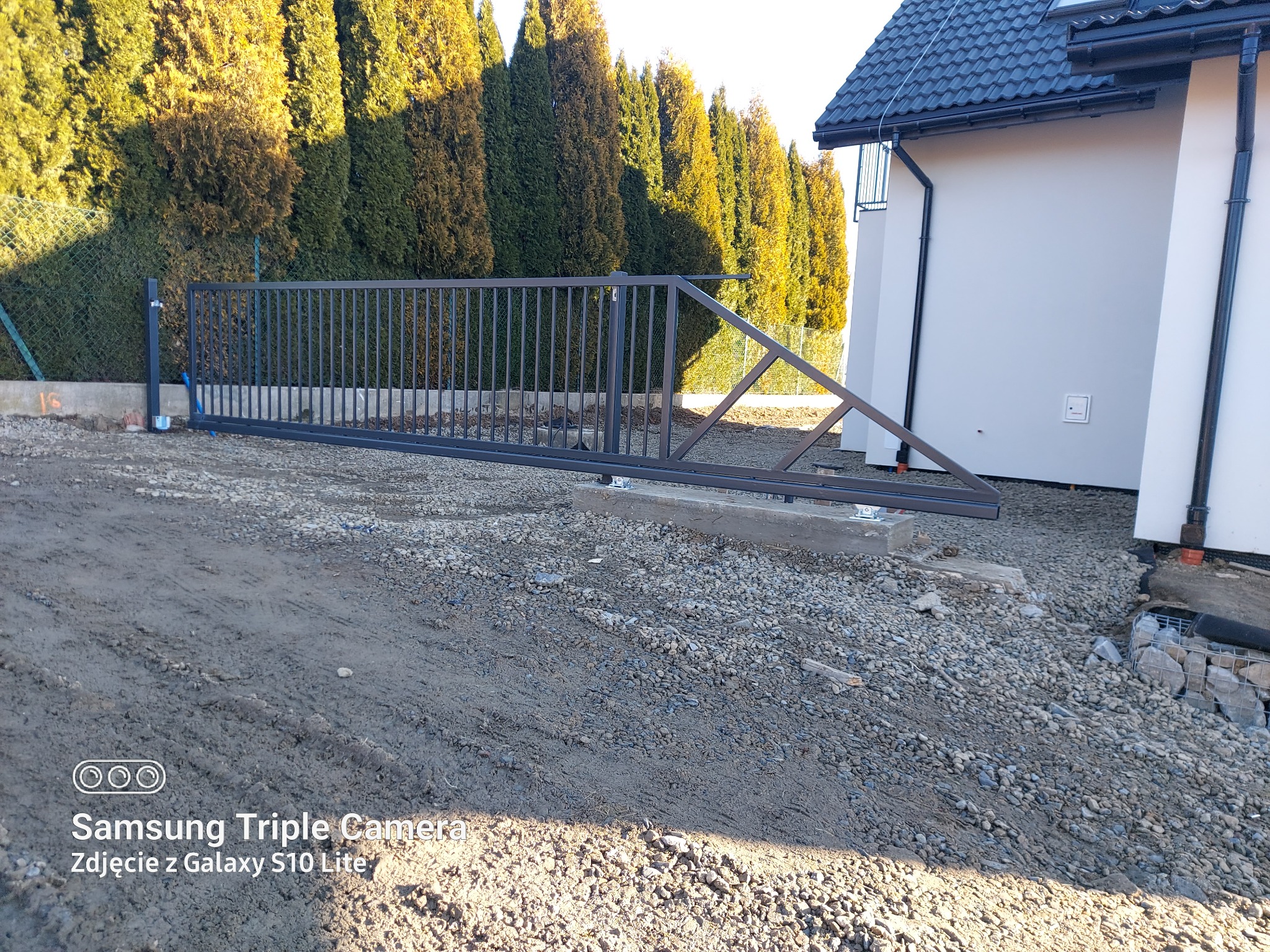 Montaż przesuwnej bramy wjazdowej z metalowymi sztachetami na betonowej wylewce, widok na dom z szarą elewacją i rząd tuj.