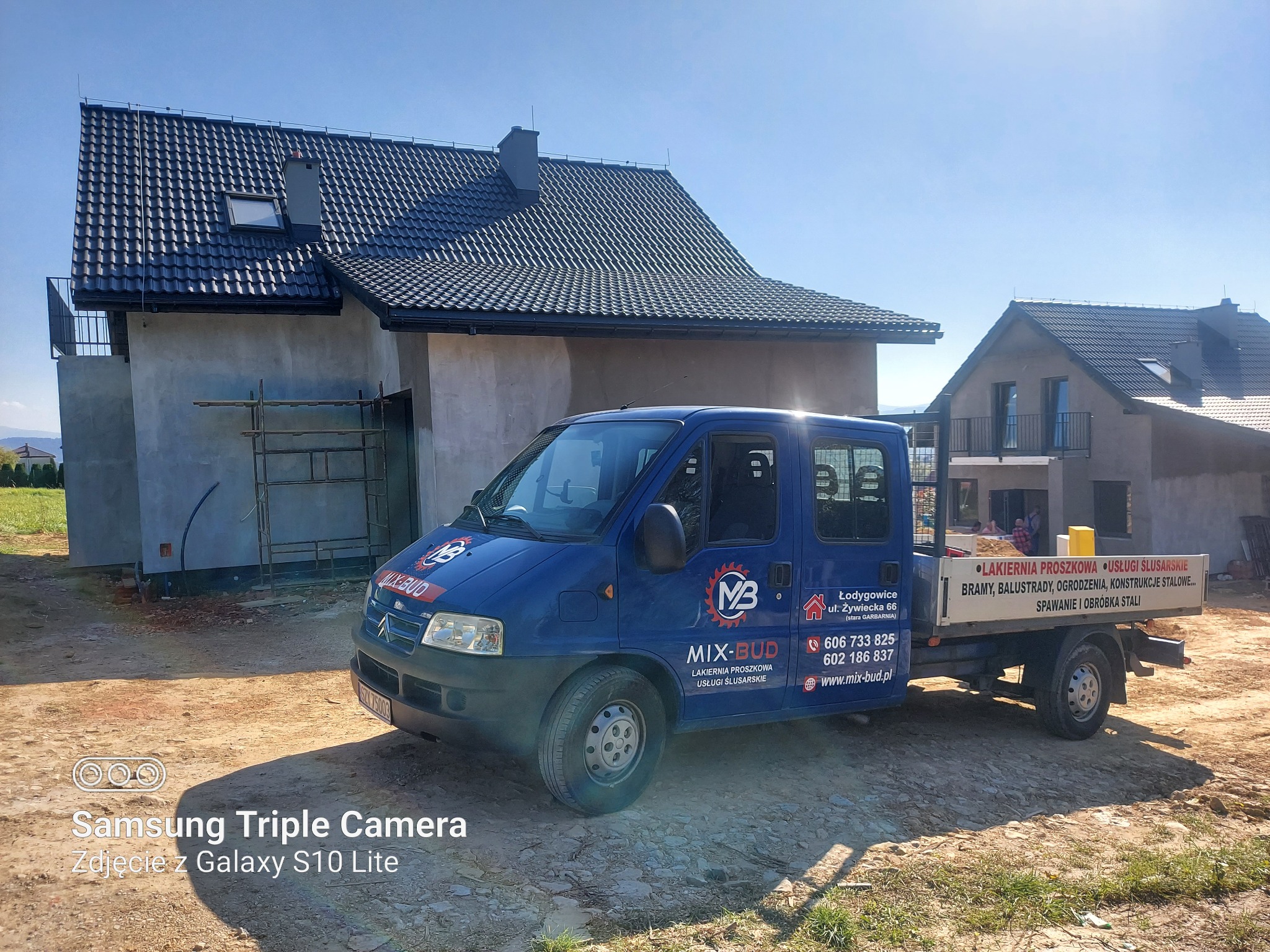 Niebieski samochód dostawczy firmy budowlanej Mix-Bud na tle dwóch budynków w stanie surowym zamkniętym, z widocznymi rusztowaniami przy jednym z nich.