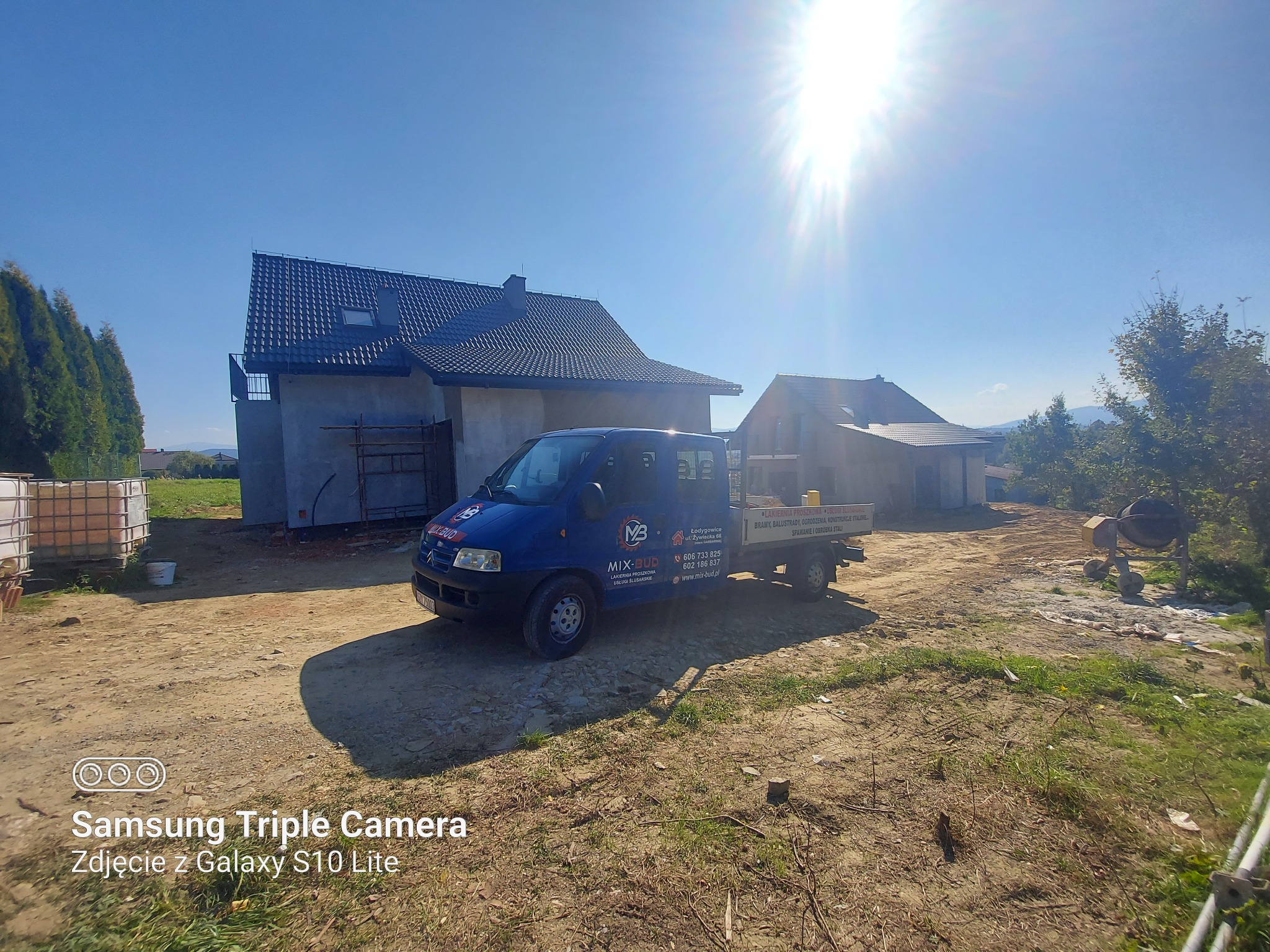 Niebieski samochód dostawczy firmy budowlanej Mix-Bud na tle dwóch budynków w trakcie budowy, widoczne rusztowania i mieszalnik do betonu.