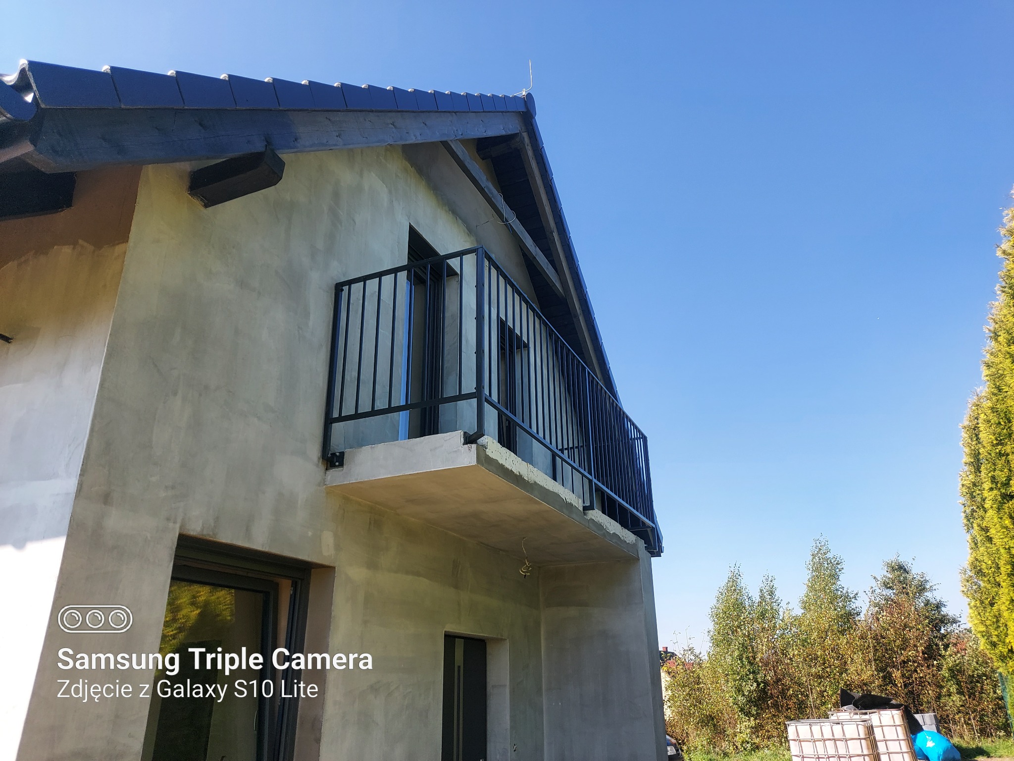 Surowy budynek z balkonem z czarną balustradą, widok z dołu na tle błękitnego nieba, ściany w trakcie wykończenia, widoczne okna i fragment dachu.