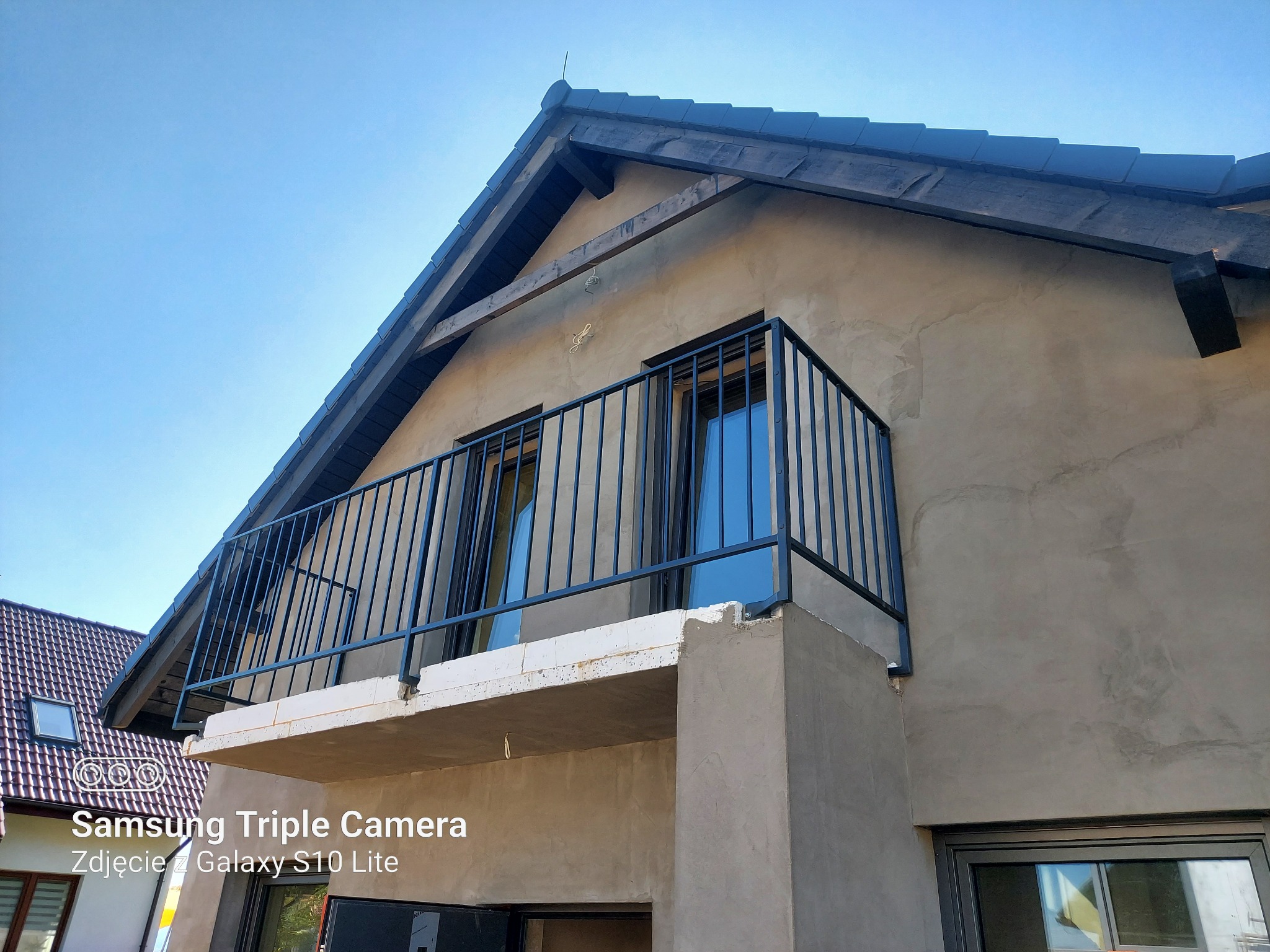 Surowy balkon z czarną, metalową balustradą w nowym budynku z szarą elewacją w trakcie prac wykończeniowych, widoczny fragment dachu i okna sąsiedniego domu.