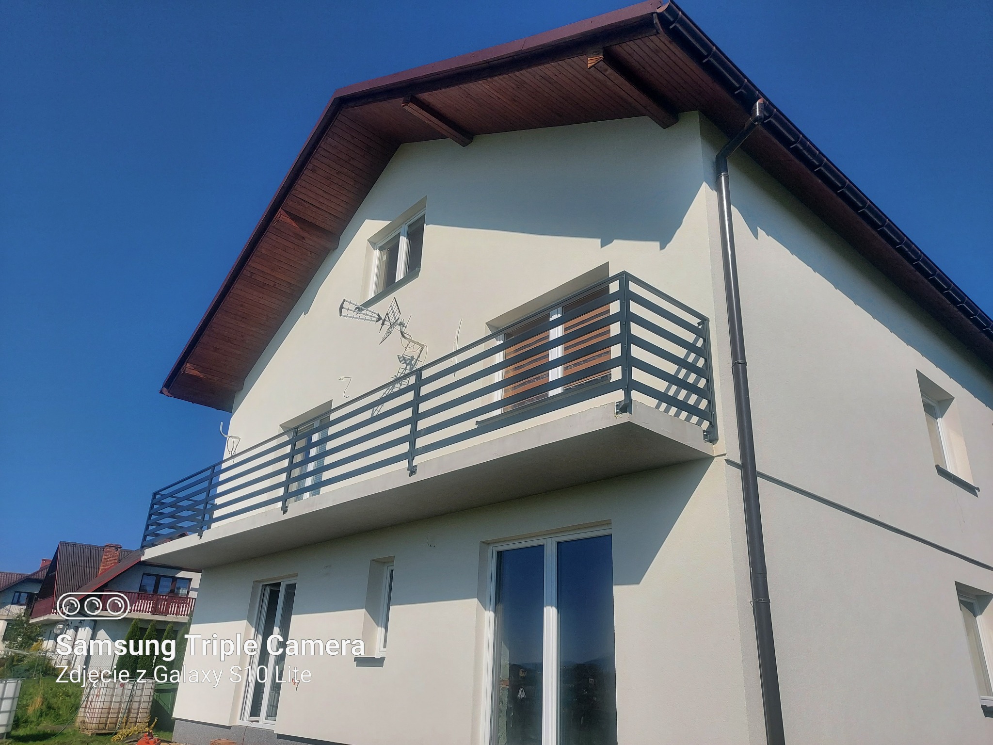 Świeżo otynkowany, dwukondygnacyjny dom z balkonami z czarną, poziomą balustradą, brązowym dachem i rynną, sfotografowany w słoneczny dzień.