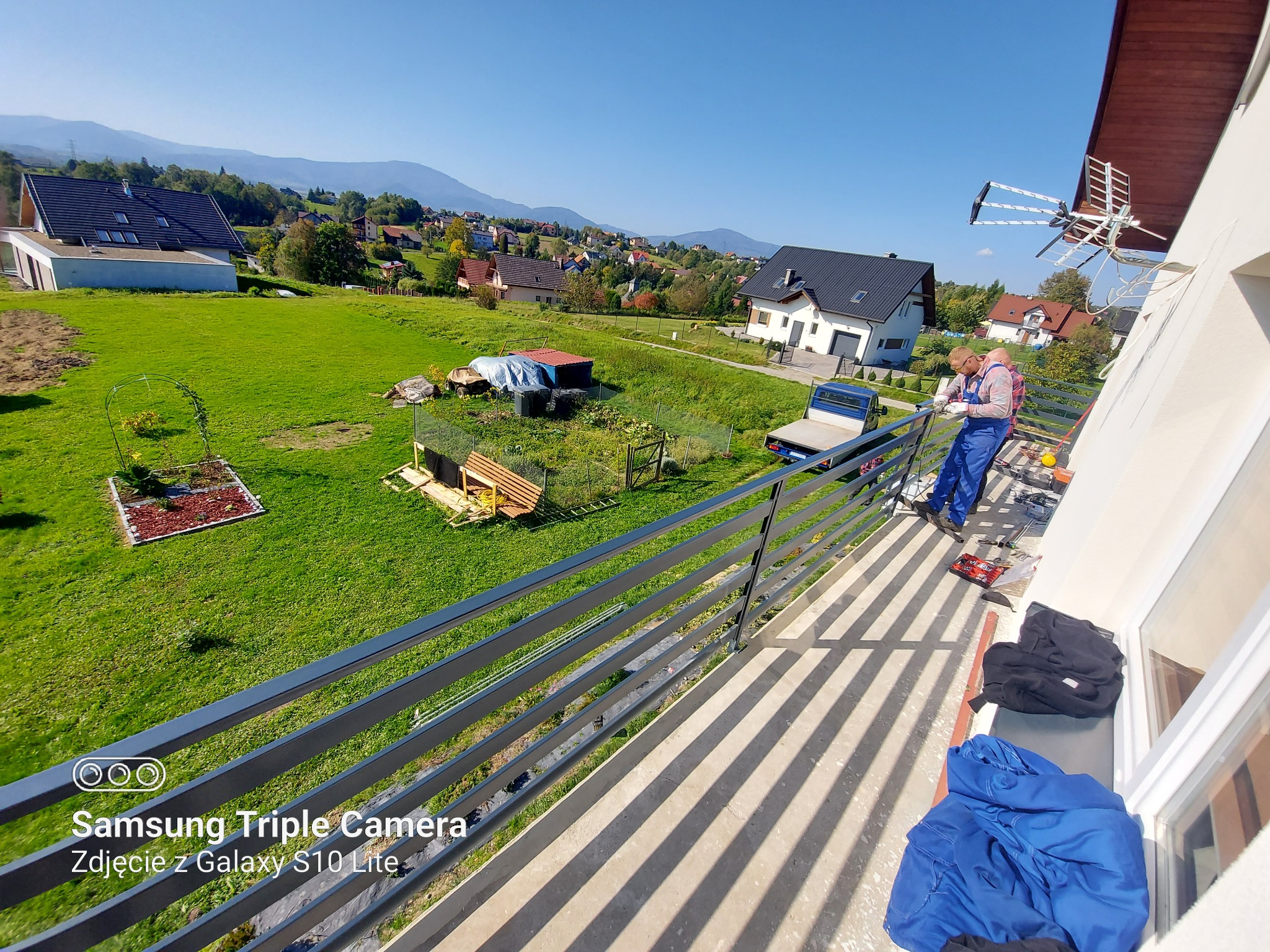 Pracownik w niebieskim kombinezonie maluje metalowe barierki balkonu na tle zielonego krajobrazu z domami i górami w oddali, widok z góry.