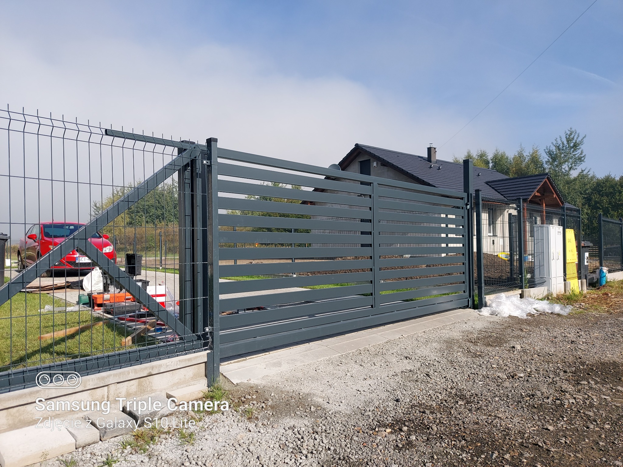 Nowoczesna, przesuwna brama posesyjna z poziomych, szarych profili, widoczna od strony zewnętrznej posesji, z fragmentem ogrodzenia z siatki i czerwonym samochodem w tle.