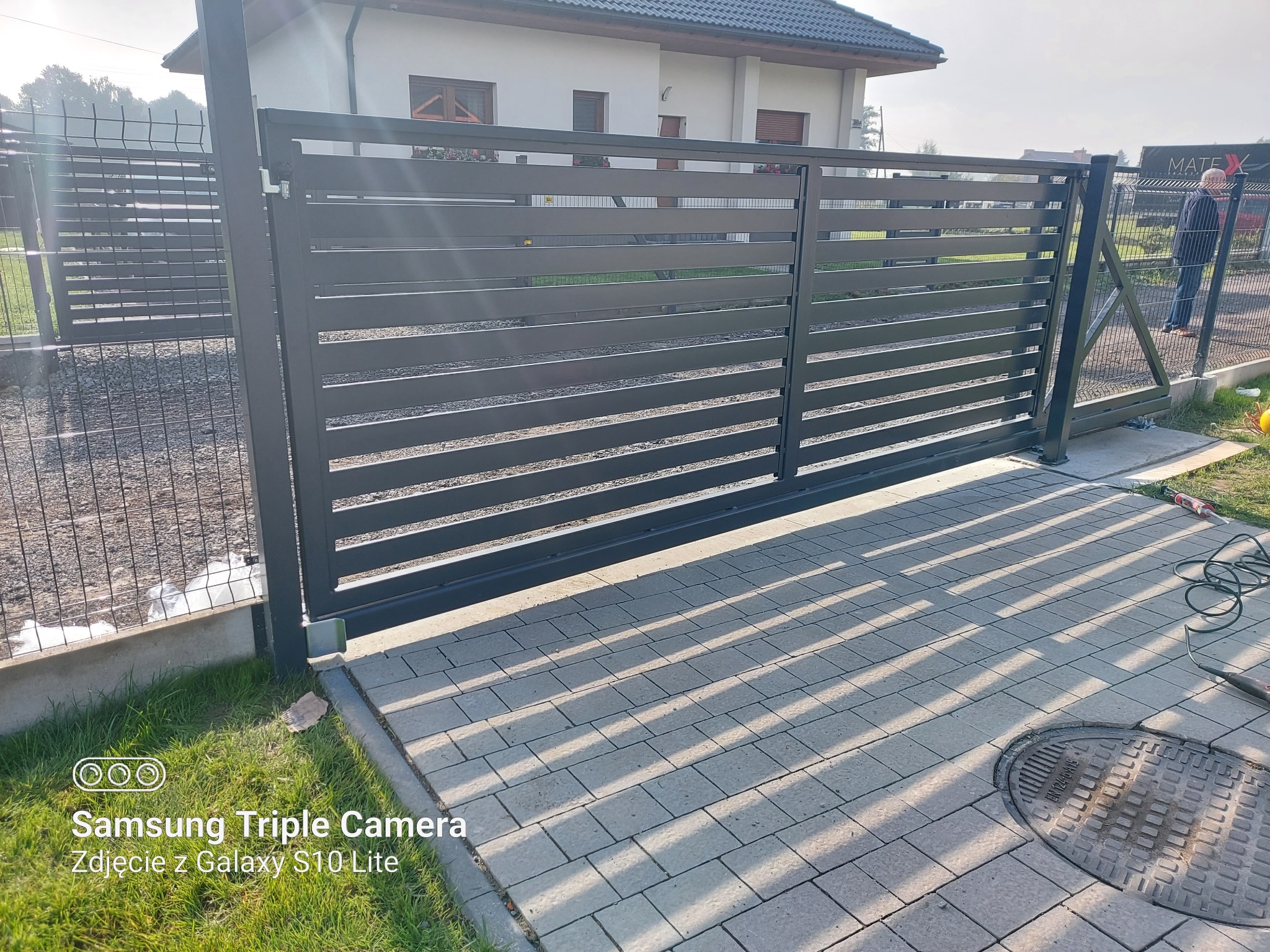 Nowoczesna, przesuwna brama posesyjna z poziomych profili, widoczna na tle domu z jasną elewacją i kostki brukowej, z narzędziami obok studzienki kanalizacyjnej.