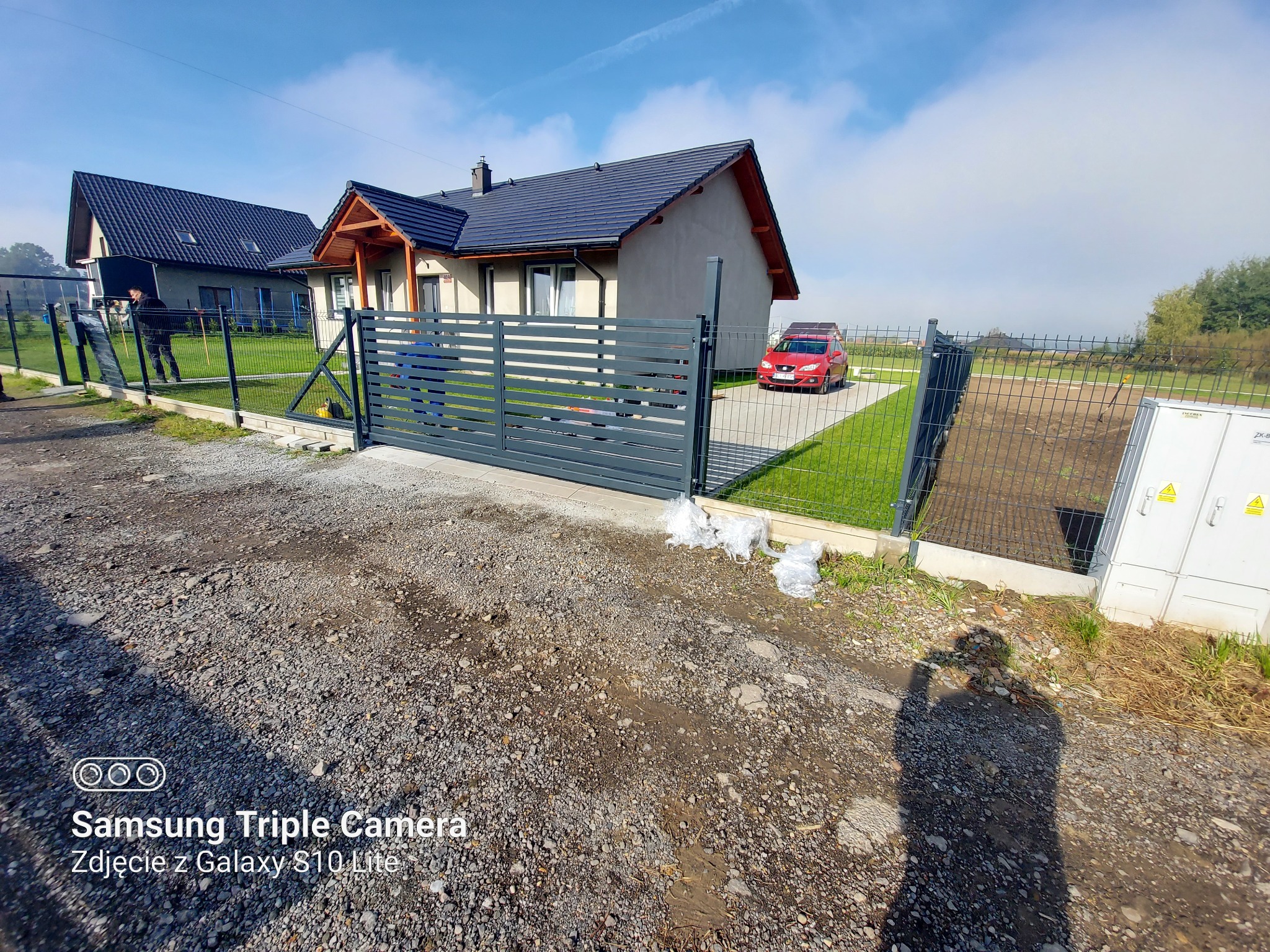 Nowoczesna posesja z szarą, przesuwną bramą wjazdową, widok na dom z ciemnym dachem i czerwonym samochodem na podjeździe, teren częściowo przygotowany pod trawnik.