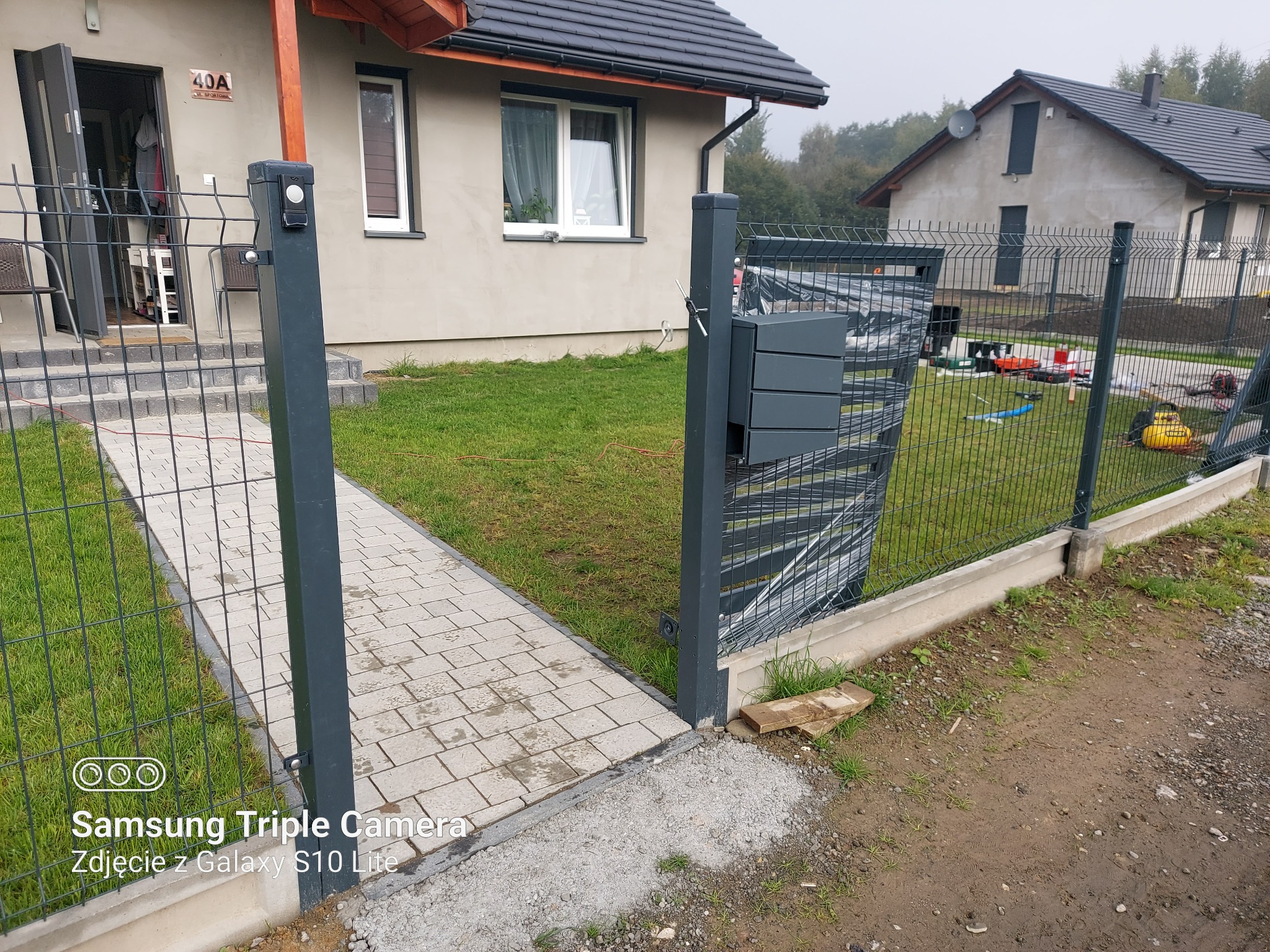 Nowoczesne ogrodzenie panelowe z furtką i skrzynką pocztową w Łodygowicach, widok na dom z kostką brukową i trawnikiem.