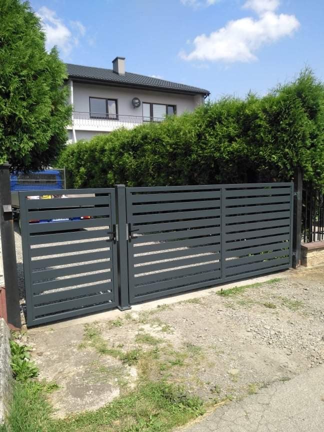 Nowoczesna, szara brama posesyjna z poziomymi szczeblami, widoczna na tle domu jednorodzinnego z zielonymi krzewami.