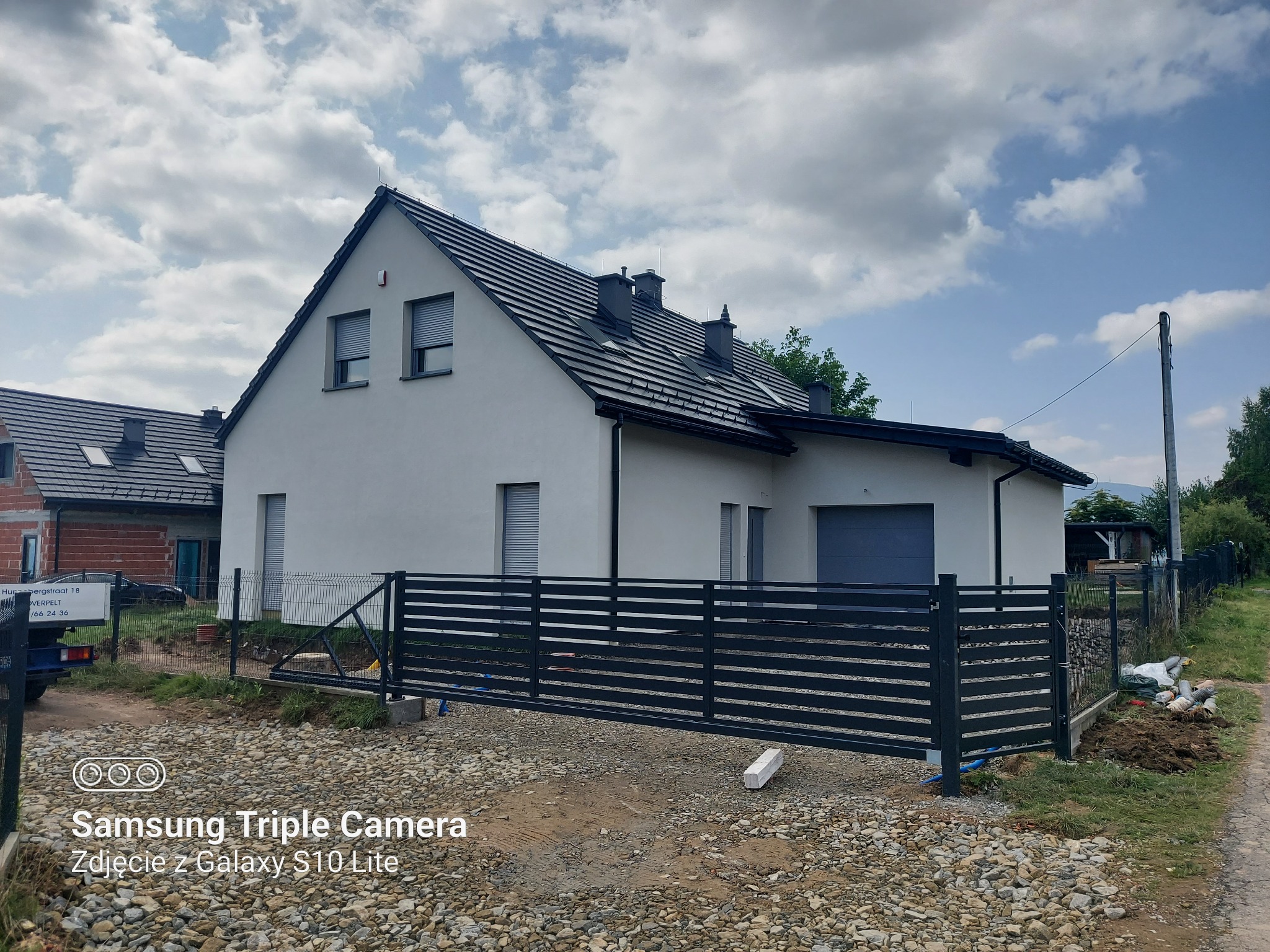 Nowoczesny dom jednorodzinny z szarą dachówką i czarną, przesuwną bramą wjazdową, widoczny z zewnątrz na tle pochmurnego nieba i pagórkowatego terenu.