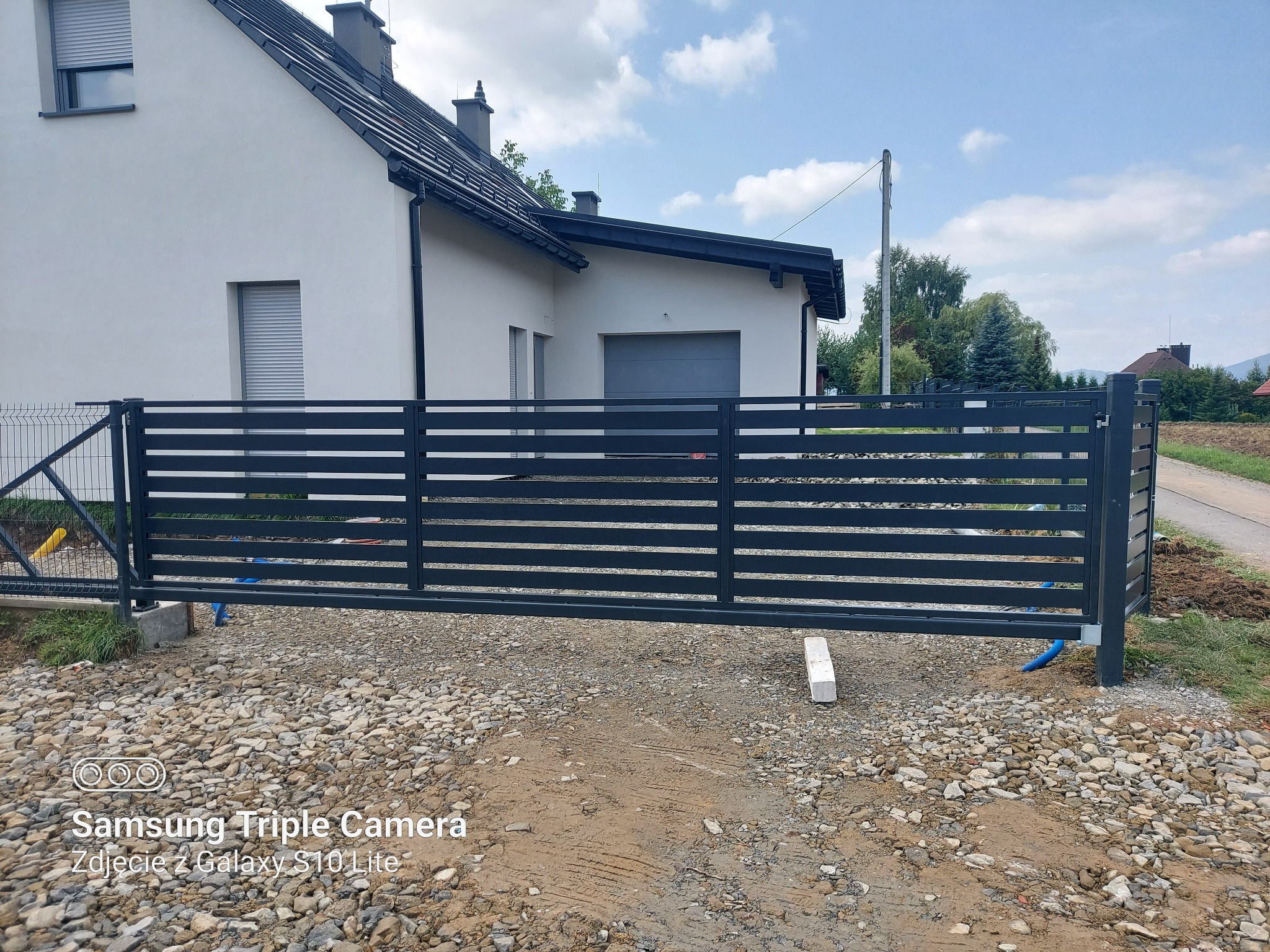 Nowoczesna, przesuwna brama wjazdowa z poziomymi panelami, zamontowana przed domem z jasną elewacją i szarą stolarką okienną, teren wokół wysypany kamieniami.