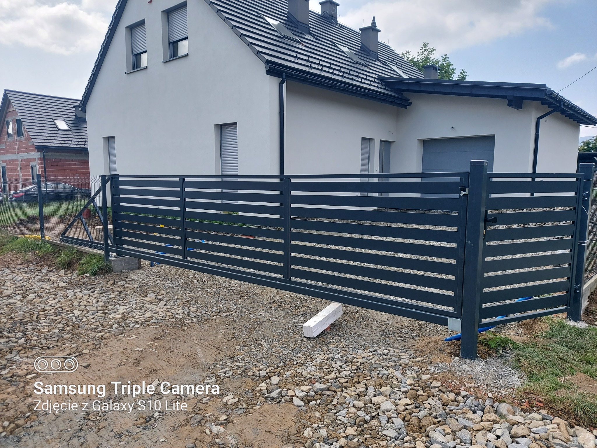 Nowoczesna, przesuwna brama wjazdowa z poziomymi panelami, pomalowana na grafitowy kolor, zamontowana przed nowo wybudowanym domem jednorodzinnym z szarą dachówką i białymi ścianami, widoczna...