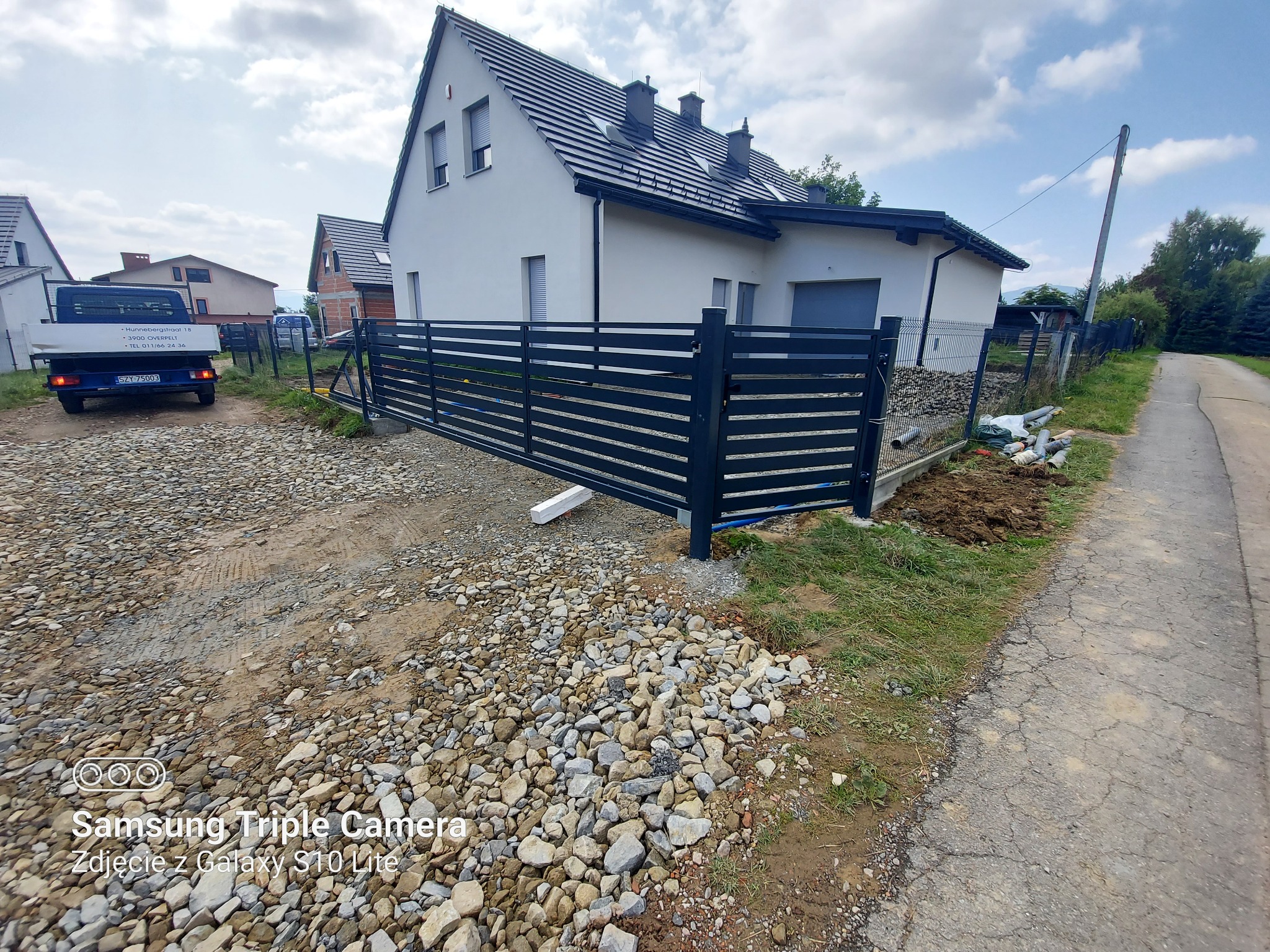 Nowoczesne, poziome ogrodzenie panelowe w kolorze antracytowym, zamontowane przed białym domem jednorodzinnym na nierównym terenie z kamieniami i piaskiem, z widocznym samochodem dostawczym w tle.