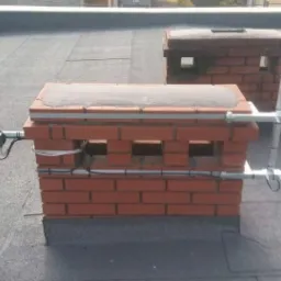 Antena satelitarna zamontowana na dachu obok komina z cegły, z widocznymi przewodami i elementami mocującymi.