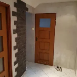 Wnętrze przedpokoju z dekoracyjną ścianą z kamienia łupanego ułożonego w nieregularny wzór, drewnianymi drzwiami z szybką i białymi płytkami podłogowymi; para butów leży przy ścianie.