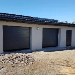 Nowo otynkowany garaż dwustanowiskowy z grafitowymi bramami segmentowymi i ciemną stolarką drzwiową, widoczny bałagan po pracach wykończeniowych.