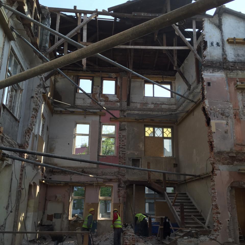 Wnętrze starej kamienicy w trakcie rozbiórki, widoczne odsłonięte ściany, drewniane elementy konstrukcyjne dachu oraz pracownicy w żółtych kaskach i kamizelkach odblaskowych.