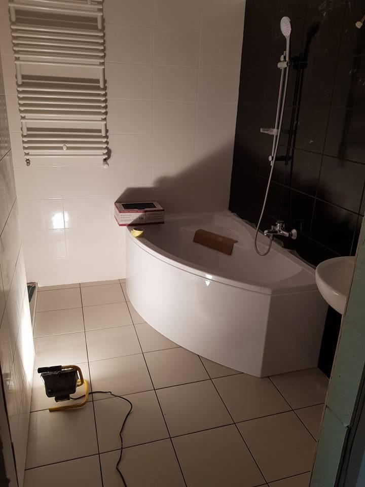 Narożna wanna w łazience po remoncie, widoczny grzejnik drabinkowy, czarne płytki na ścianie, lampa budowlana oświetlająca podłogę z jasnych płytek, pudełko z akcesoriami na wannie.