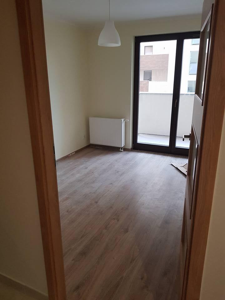 Pusty pokój po remoncie z panelami podłogowymi w odcieniach brązu, białym grzejnikiem, lampą wiszącą i drzwiami balkonowymi z widokiem na budynek mieszkalny. Widok zza otwartych drewnianych drzwi.