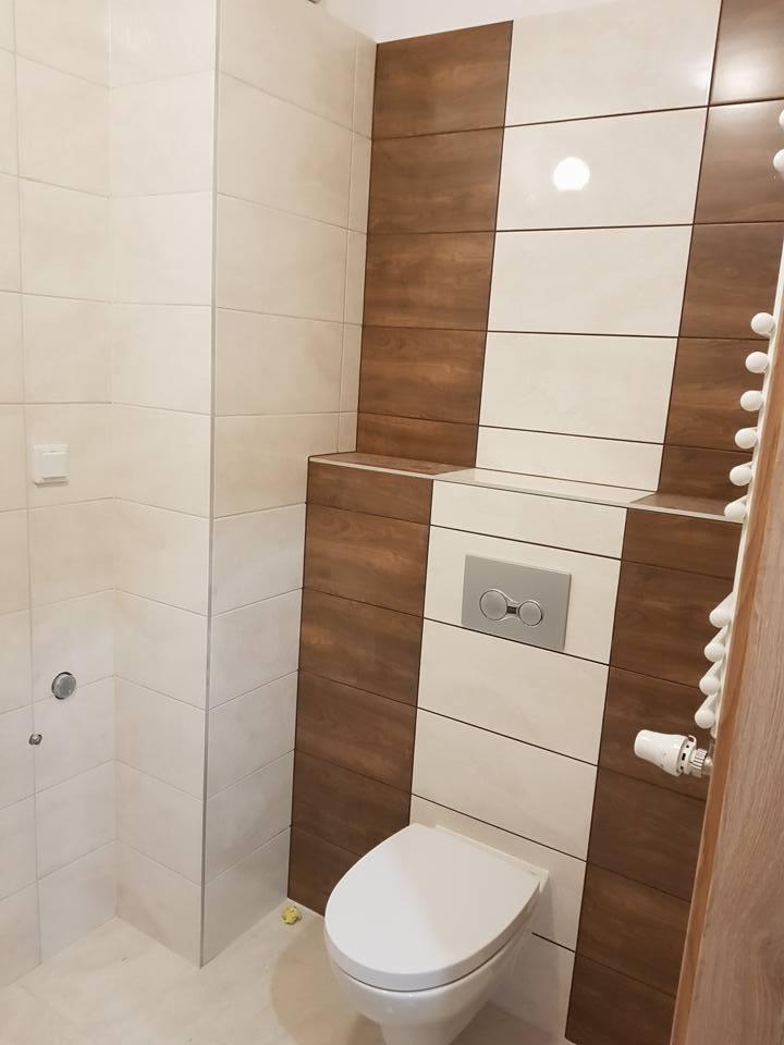 Nowoczesna łazienka z białymi i drewnopodobnymi płytkami ułożonymi w pionowe pasy, wisząca toaleta z chromowanym przyciskiem spłukiwania, grzejnik drabinkowy po prawej stronie.