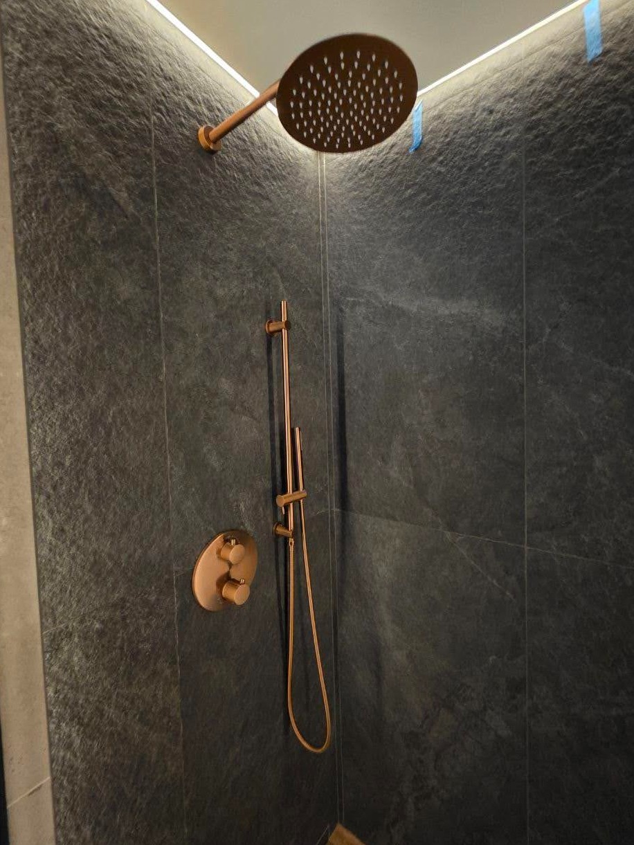 Nowoczesna łazienka z ciemnymi płytkami imitującymi kamień i miedzianą armaturą: deszczownica, bateria i słuchawka prysznicowa. Oświetlenie LED podkreśla surowy charakter.