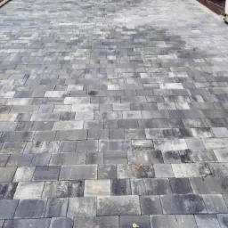 Ułożona, szara kostka brukowa o nieregularnych kształtach, widoczna z perspektywy chodnika, z ziemią po lewej stronie i fragmentem ściany budynku po prawej.