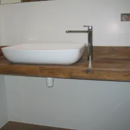 Umywalka nablatowa o kwadratowym kształcie z chromowaną baterią na drewnianym blacie z płytek imitujących drewno. Widoczne białe płytki na ścianach, wystający kabel elektryczny i podłączenie...