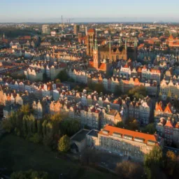 Panorama Gdańska z lotu ptaka, ukazująca gęstą zabudowę Starego Miasta z charakterystycznymi czerwonymi dachami i wieżami kościołów w promieniach porannego słońca.