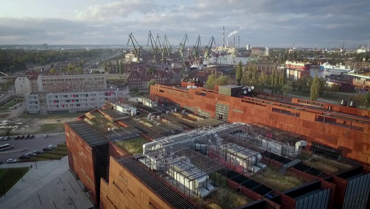 Szeroki kadr z lotu ptaka na budynek pokryty rdzawym metalem z zielonymi dachami, w tle widok na port z żurawiami i kominami, oraz zabudowania miejskie.