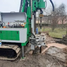 Wiertnica na gąsienicach w trakcie pracy, woda wypływa z odwiertu, błotniste podłoże.