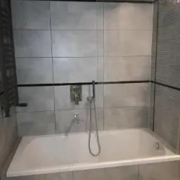 Szara łazienka z wanną obłożoną płytkami, baterią podtynkową i szklanym panelem prysznicowym.