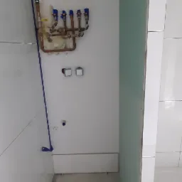 Wnętrze łazienki w trakcie prac: widoczne rury miedziane z niebieskimi zaworami, białe płytki na ścianach, częściowo położona płyta karton-gips oraz niezakończona wylewka podłogowa.