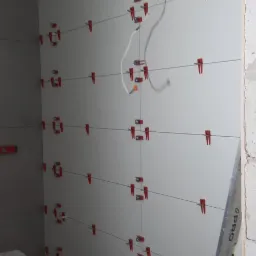 Ściana w trakcie układania płytek, widoczne czerwone klipsy poziomujące, białe przewody elektryczne wystające ze ściany, poziomica oparta o ścianę, drabina w rogu.