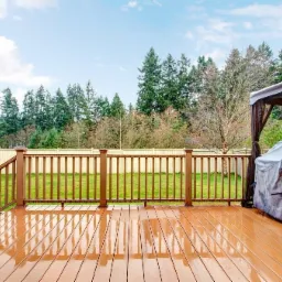 Błyszczący drewniany taras z brązową balustradą, widok na trawnik, jasny płot i zielone drzewa w tle, zadaszony grill po prawej stronie.