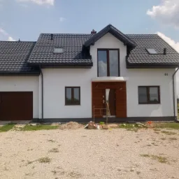 Nowoczesny dom jednorodzinny z białą elewacją i grafitową dachówką, widoczny fragment terenu przed domem z piaskiem i narzędziami malarskimi, sugerujący prace wykończeniowe.