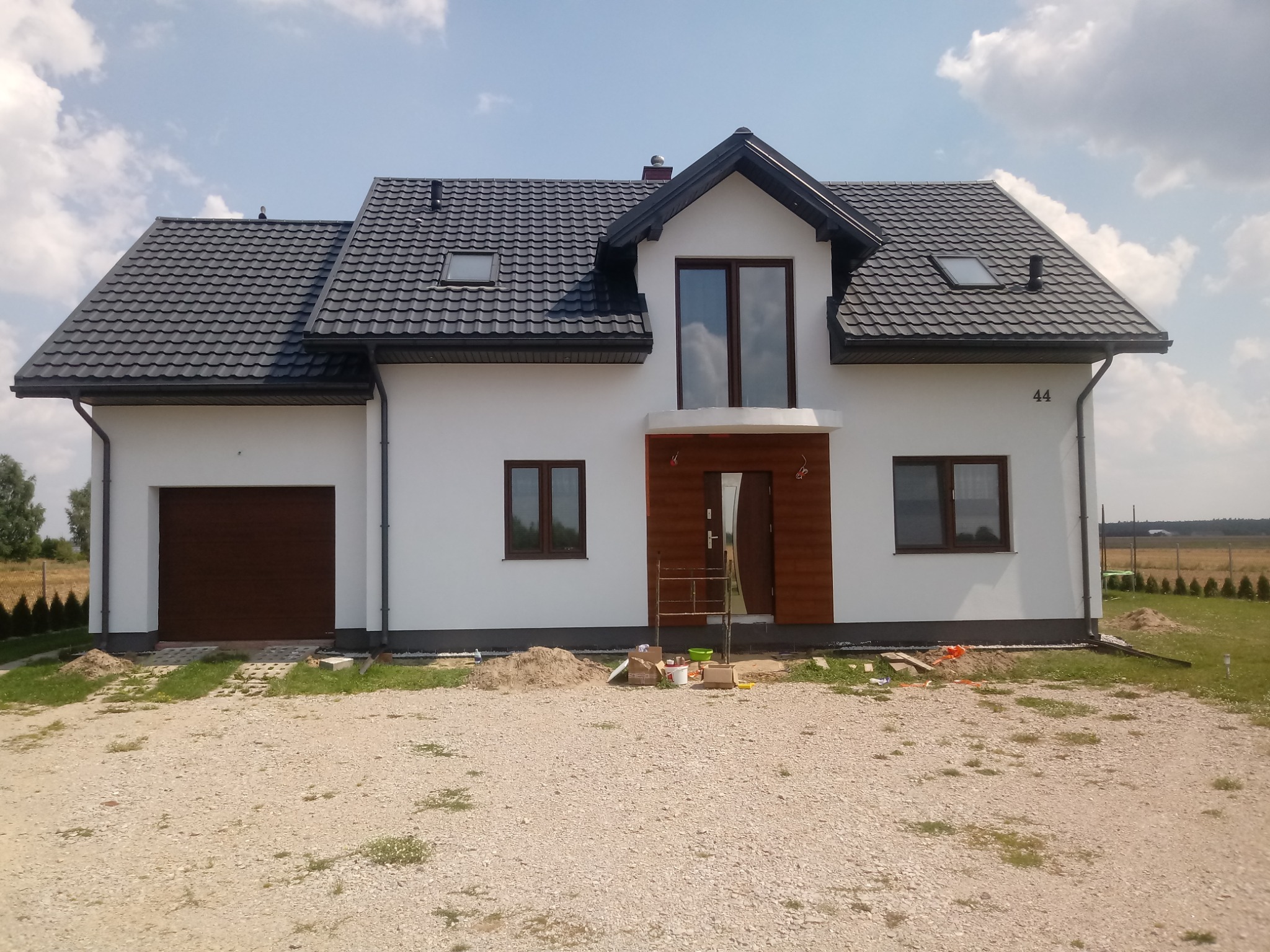 Nowoczesny dom jednorodzinny z białą elewacją i grafitową dachówką, widoczny fragment terenu przed domem z piaskiem i narzędziami malarskimi, sugerujący prace wykończeniowe.