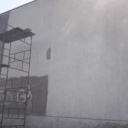Fragment budynku z widocznymi śladami po usunięciu graffiti i częściowo odnowioną elewacją, rusztowanie z myjką ciśnieniową w lewym dolnym rogu.