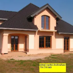 Nowo wybudowany, dwukondygnacyjny dom z ciemnobrązową dachówką i oknami w drewnianych ramach, z elementami elewacji w odcieniach beżu i kremu, widoczne ślady prac budowlanych wokół domu.