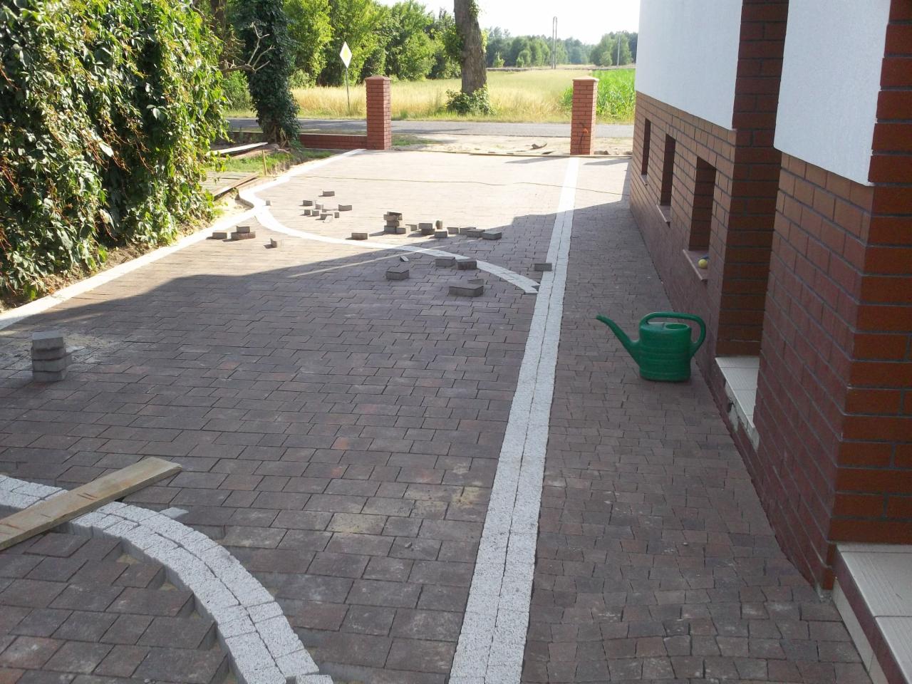 Układanie kostki brukowej na podjeździe do domu, widoczne niedokończone fragmenty obrzeży i porozrzucane kostki, zielona konewka przy ścianie budynku.