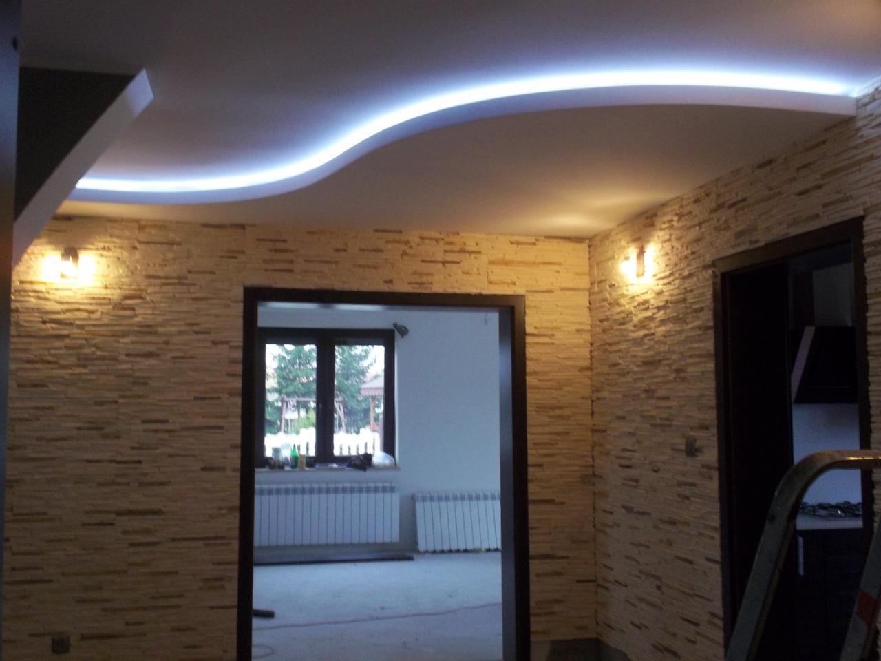 Wykończone wnętrze: ściany wyłożone dekoracyjnym kamieniem w odcieniach beżu i brązu, podświetlany sufit podwieszany z falistą linią LED, widoczne dwie pary drzwi z ciemną stolarką.