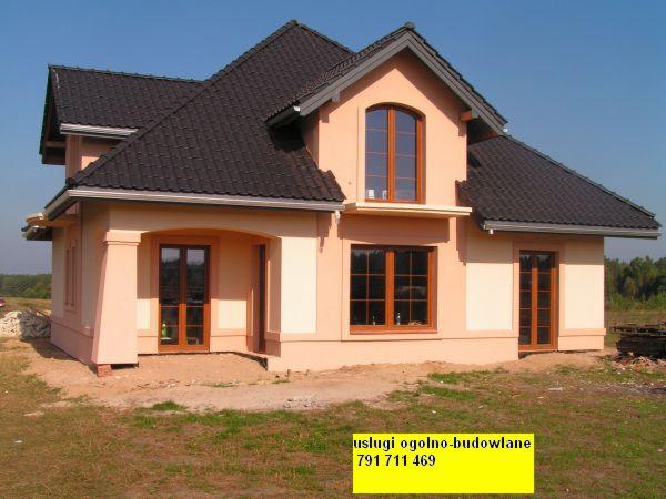 Nowo wybudowany, dwukondygnacyjny dom z ciemnobrązową dachówką i oknami w drewnianych ramach, z elementami elewacji w odcieniach beżu i kremu, widoczne ślady prac budowlanych wokół domu.