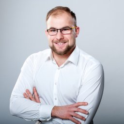 SEOmotive Agencja Interaktywna - Oficjalny Partner Google - Portret uśmiechniętego mężczyzny w okularach i białej koszuli, z założonymi rękami na tle szarego studia. Profesjonalny wizerunek przedsiębiorcy.