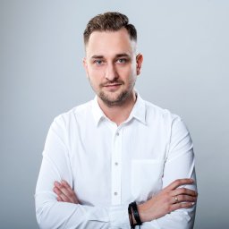 SEOmotive Agencja Interaktywna - Oficjalny Partner Google - Portret biznesowy mężczyzny w białej koszuli z założonymi rękami na tle szarego studia. Widoczny pierścionek i czarna bransoletka na nadgarstku.