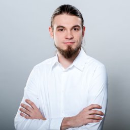 SEOmotive Agencja Interaktywna - Oficjalny Partner Google - Portret mężczyzny z brodą i włosami związanymi w kucyk, ubranego w białą koszulę, z założonymi rękami na tle szarego studia.